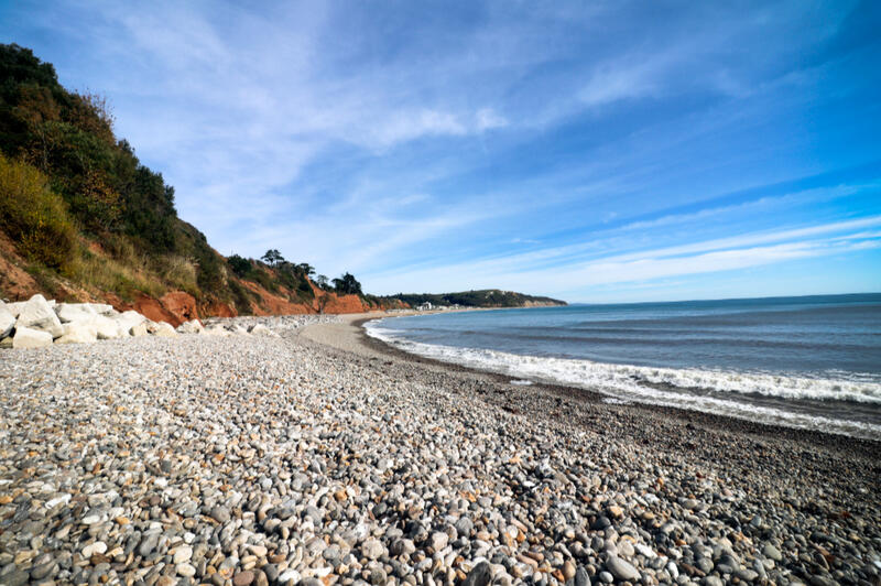 Pebble Shoreline at Seaton Beach, Devon Pebble beach and calm sea at Seaton in Devon, England, with red cliffs and a bright blue sky stretching along the coastline.